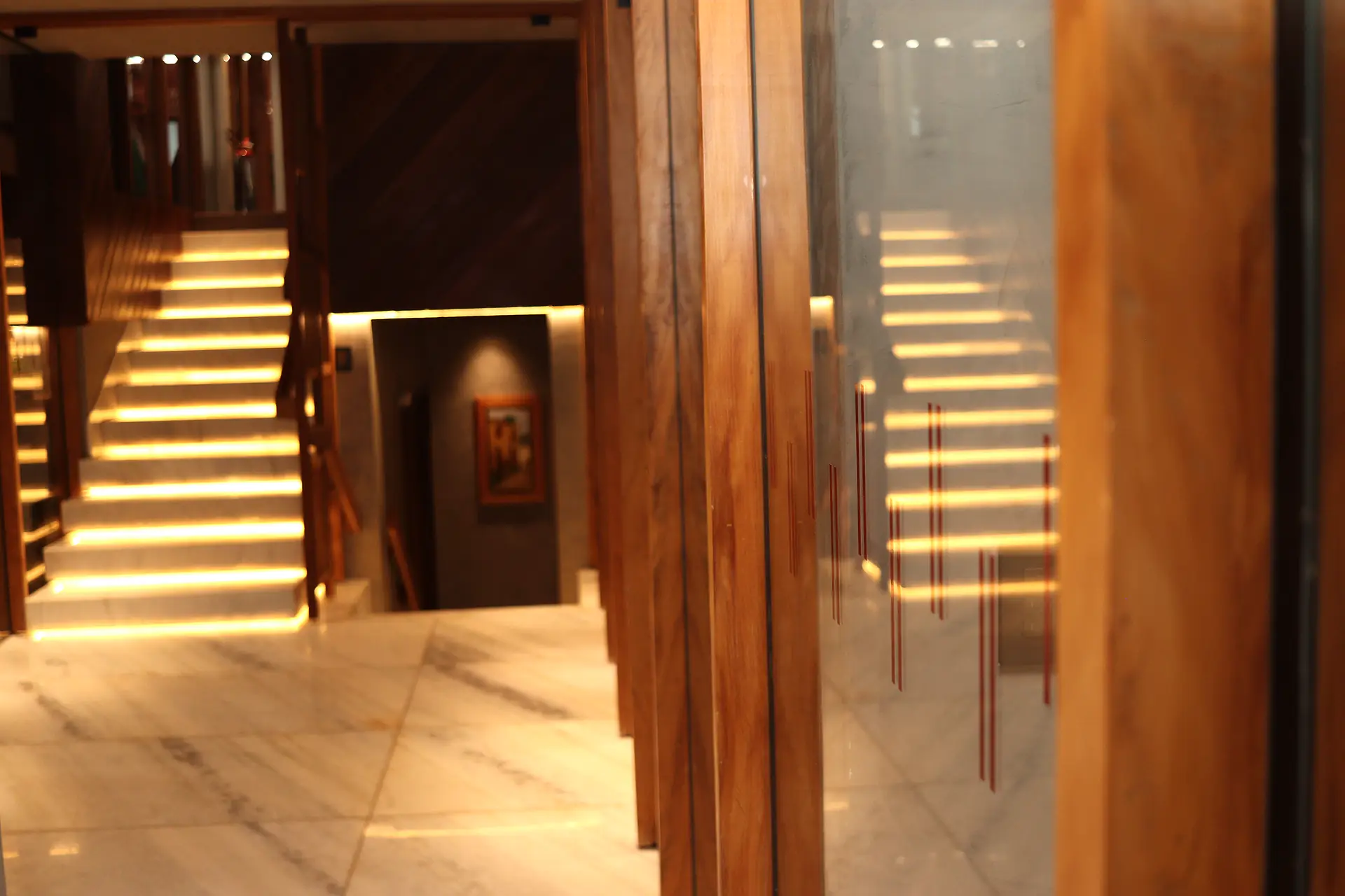 Escada de mármore com degraus iluminados por fita de LED, emoldurada por ripas de madeira e reflexo no vidro do corredor
