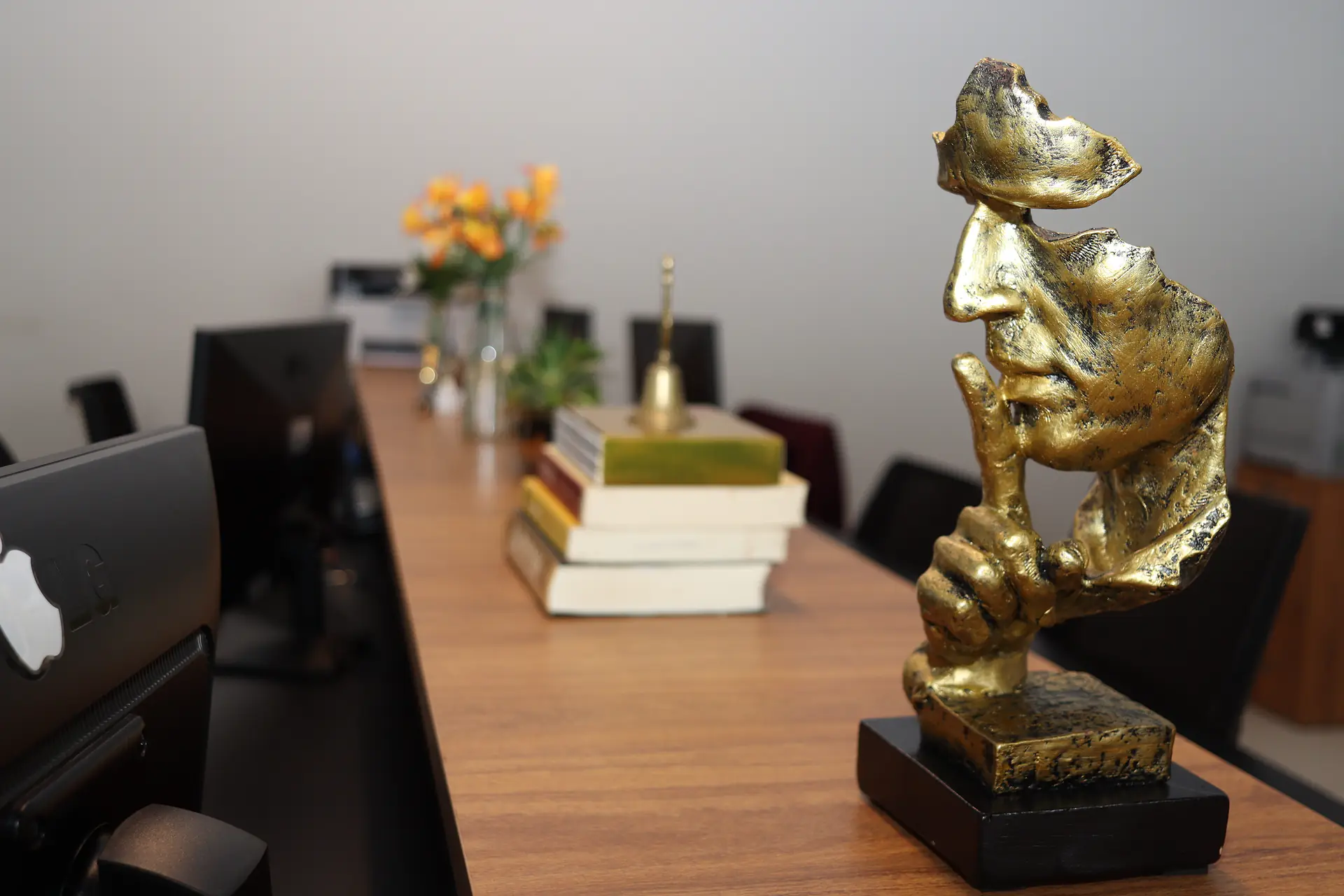 Escultura dourada de rosto com o dedo sobre os lábios, símbolo de sigilo, sobre bancada de madeira com livros empilhados e flores laranja ao fundo