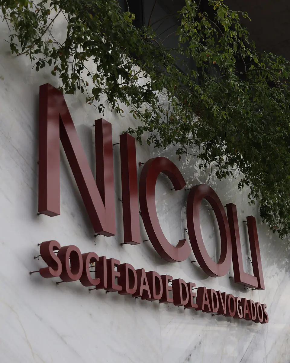 Letreiro "Nicoli Sociedade de Advogados" em letras bordô tridimensionais sobre a fachada de mármore branco, com folhagens pendentes