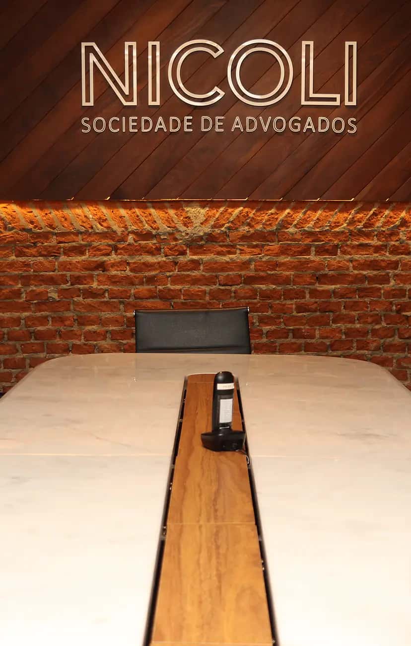 Sala de reuniões da Nicoli Sociedade de Advogados com letreiro "Nicoli Sociedade de Advogados" em aço escovado sobre painel de madeira em diagonal, parede de tijolos com iluminação rasante e mesa de mármore branco com cadeiras executivas pretas