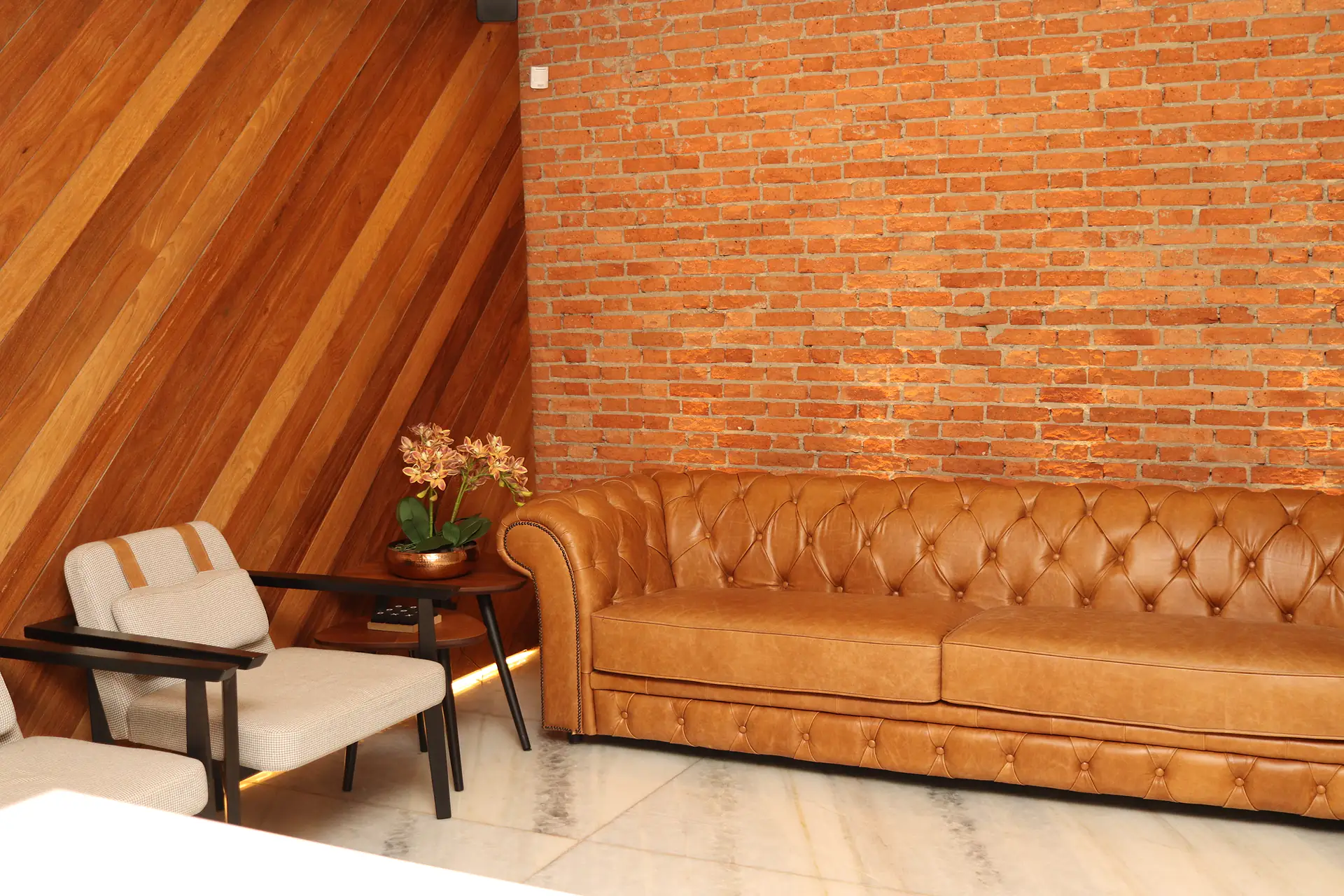 Sala de espera da Nicoli Sociedade de Advogados com sofá chesterfield de couro caramelo, poltronas claras, parede de tijolos aparentes e revestimento de madeira em diagonal