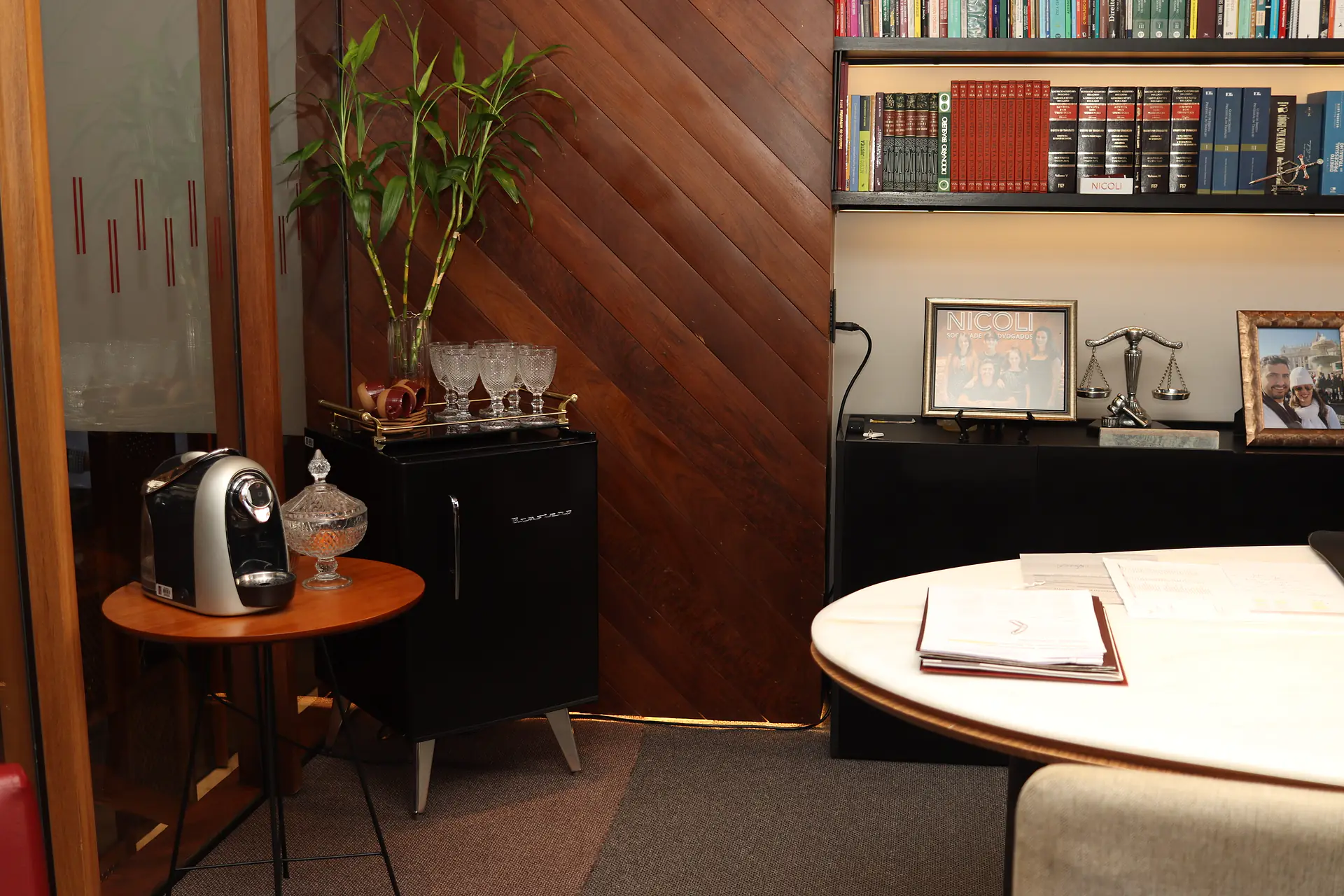 Canto da sala do sócio com máquina de café, taças de cristal, bambu da sorte, parede de madeira em diagonal, estante de livros e mesa de mármore